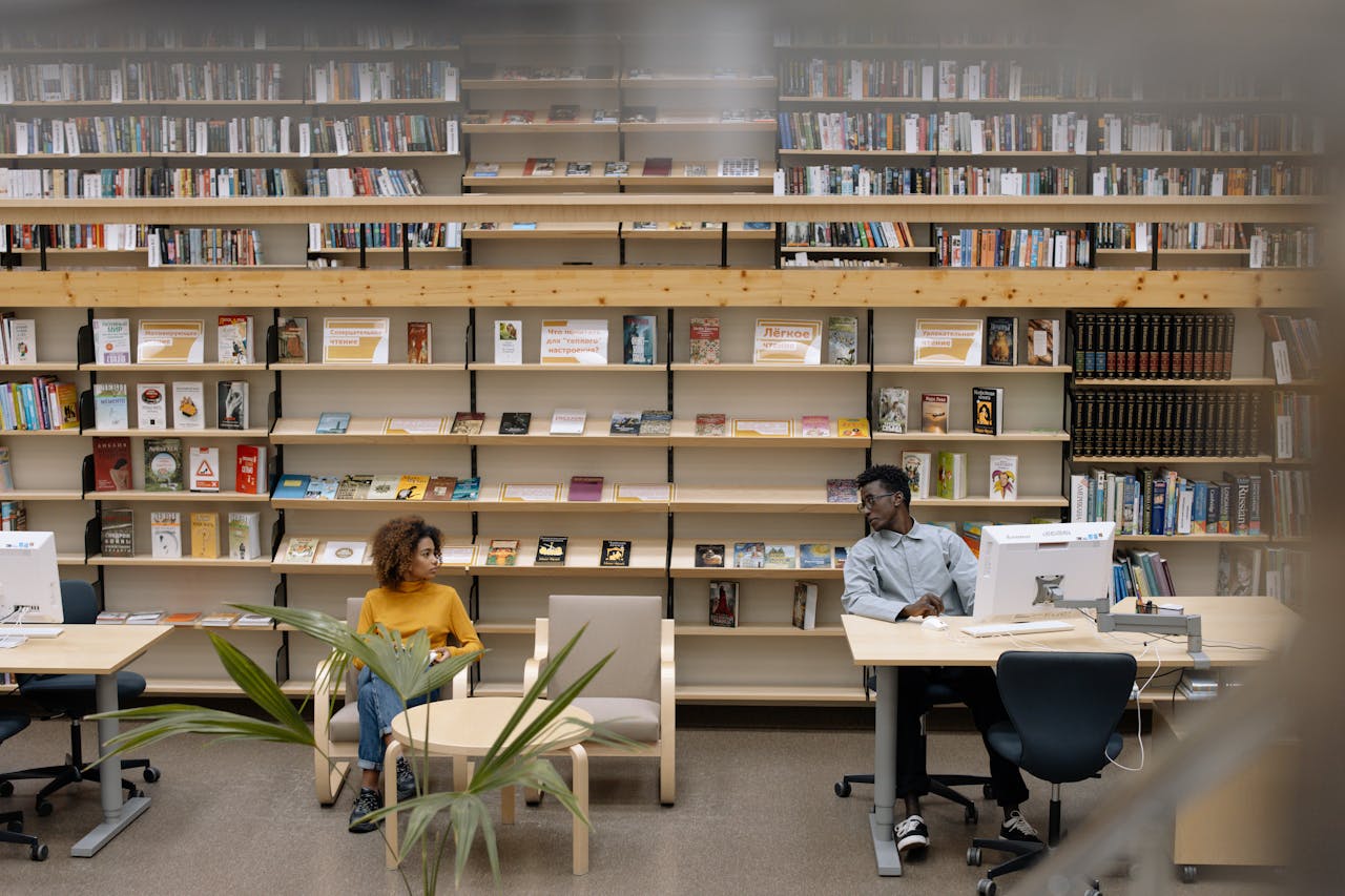 Two adults engaged in reading activities in a public library with abundant bookshelves.